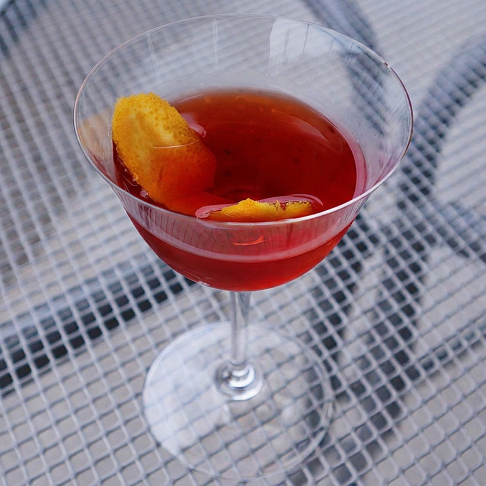 Red cocktail in a stemmed glass garnished with orange peel, resting on a patterned outdoor table.